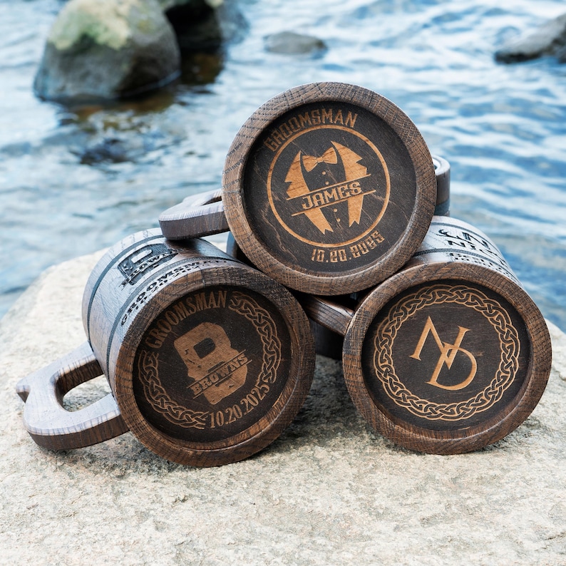 May include: Several wooden beer tankards with dark brown detailing, each engraved with custom text. The text includes names, dates, and the word "Groomsman." The mugs are displayed on a rock near water, suggesting an outdoor setting.