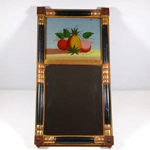 May include: Rectangular mirror with a black and gold frame. The top section features a painted still life of a pineapple, two apples, and green leaves against a blue background. The lower section is a reflective surface.