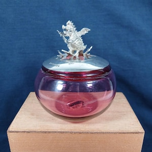 May include: A round, translucent purple glass jar with a polished silver-tone metal lid. The lid is topped with a detailed, three-dimensional silver-tone metal dragon figurine with spread wings, perched atop a decorative, star-shaped metal base.