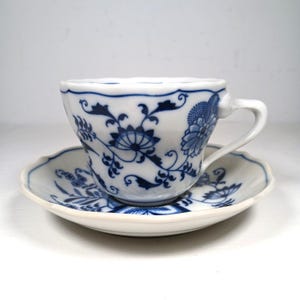 Könnte beinhalten: Weißes Porzellan-Tee-Set mit blauem Blumenmuster. Die Tasse hat einen geschwungenen Henkel und einen gewellten Rand. Die Untertasse hat ebenfalls einen gewellten Rand, und beide Teile sind mit detaillierten blauen Blumenmustern verziert.