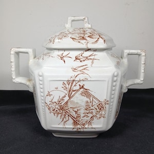 以下が含まれることがあります： 鳥、植物、風景の茶色のイラストが描かれた、蓋付きの白いセラミックシュガーボウル。 ボウルには2つの正方形のハンドルと正方形の蓋が付いています。 ヴィンテージデザイン。