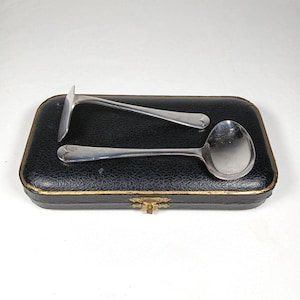 May include: Silver serving spoon and a silver server tool on a black leatherette box with a gold trim. The spoon has a round bowl, and the server has a flat, angled blade. The box is rectangular with a gold clasp.
