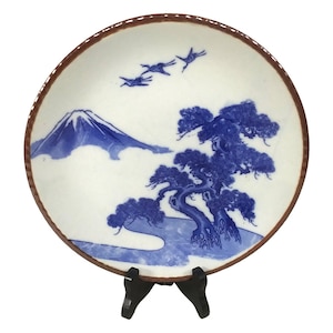 Könnte beinhalten: Ein blauer und weißer Keramikteller mit einer japanischen Berglandschaft und einem Baum. Der Teller hat einen braunen Rand und steht auf einem schwarzen Ständer.