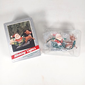 May include: A Rudolph the Red-Nosed Reindeer blinking lights ornament set featuring a Santa in a green sleigh pulled by a reindeer. The set includes a cardboard box with the text Blinking Lights and a clear plastic tray containing the ornament.