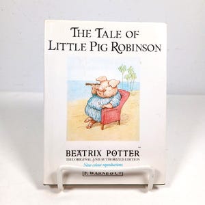 Puede incluir: Un libro de tapa dura titulado The Tale of Little Pig Robinson de Beatrix Potter. La cubierta blanca presenta una ilustración a color de un cerdo con camisa azul sentado en una silla roja en una playa, sosteniendo un telescopio. El texto incluye el nombre de la autora y la editorial F. Warne & Co.