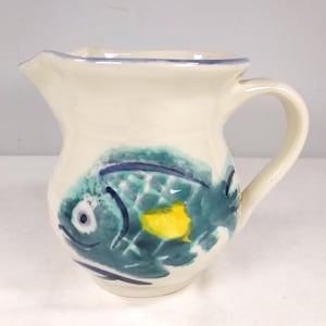 May include: A white ceramic pitcher with a blue and green fish design. The fish has a yellow spot on its side.