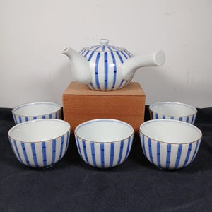 May include: A white ceramic tea set with blue vertical stripe design. The set includes a teapot with a lid and a handle, and five matching tea cups. The tea set is displayed on a wooden box.