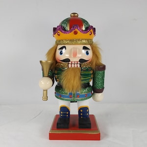 Könnte beinhalten: Eine Holz-Nussknacker-Figur mit grüner und goldener Uniform, einer rot-violetten Krone und einer goldenen Trompete. Der Nussknacker hat ein weißes Gesicht mit einem schwarzen Schnurrbart und Bart.