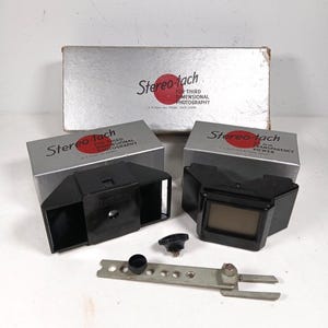 Op de afbeelding: Twee zwarte Stereo-tach 3D-fotografie-opzetstukken met zilveren dozen met het label Stereo-tach for third dimensional photography. Inclusief een metalen montagebeugel met ronde gaten, een kleine zwarte knop en een zwarte dop voor camera-apparatuur.