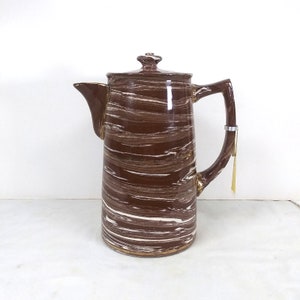 Può includere: Una caffettiera in ceramica marrone e bianca con un motivo a spirale. La caffettiera ha un bordo dorato intorno alla parte superiore e inferiore.