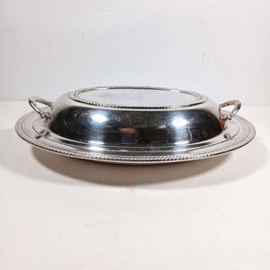 May include: A polished silver-tone oval serving dish with a domed lid and two side handles. The dish features a decorative rope-style border along the rim of the lid and the base, with a smooth, reflective metallic surface throughout the piece.