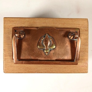 Puede incluir: Una caja rectangular de madera con una placa central de cobre que presenta un emblema en relieve con forma de flor o hoja estilizada. La placa cuenta con un asa de cobre en forma de U, sujeta por dos anillas, con un acabado metálico.