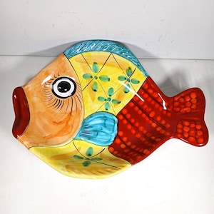 May include: A ceramic fish-shaped serving dish featuring a hand-painted design with a large black eye, an orange face, a yellow body with green floral accents, a blue fin, and a deep red tail section decorated with a repeating dot pattern.