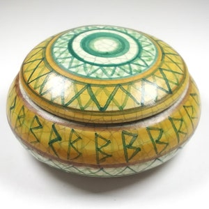 Puede incluir: Una caja redonda de cerámica para baratijas con tapa. La caja está decorada con patrones geométricos verdes, amarillos y marrones. La tapa tiene un diseño circular verde y blanco en el centro.