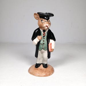 May include: A ceramic figurine of a rabbit dressed in academic attire, including a black cap and gown, a green vest, and a striped tie. The rabbit holds a small red book and stands on a brown base. The figurine has a detailed, hand-painted appearance.