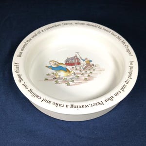 May include: White ceramic bowl featuring a colorful illustration of Peter Rabbit running from Mr. McGregor. The bowl has text around the rim, and the scene depicts a garden setting with a red building.