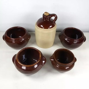 Puede incluir: Cinco cuencos de cerámica marrón con una jarra de cerámica marrón y beige en el centro. Los cuencos tienen bordes redondeados y un labio ligeramente acampanado. La jarra tiene un tapón de corcho y un asa.