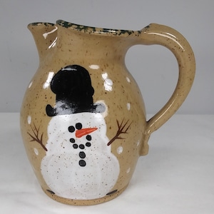 May include: A brown ceramic pitcher with a snowman design. The snowman is wearing a black hat and has a carrot nose and black eyes. The pitcher has a speckled brown glaze with white dots.