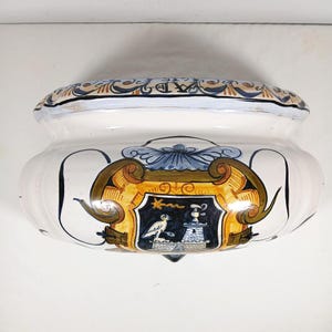 Puede incluir: Una maceta de cerámica blanca con un borde decorativo y un diseño detallado de escudo de armas. La maceta presenta un escudo con un pájaro, una torre y una estrella, acentuado con detalles azules, dorados y marrones. El diseño general es ornamentado y clásico.