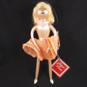 May include: A glass ornament of a ballerina wearing a peach-colored dress with a pink ribbon and a pink ruffled collar. The ornament has blonde hair and a smiling face. The ornament has a tag that says "Beau  Arts".