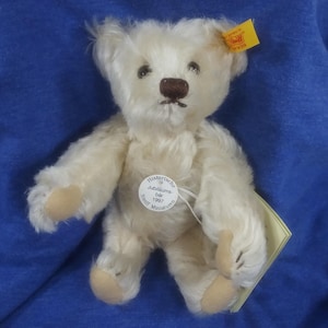 May include: A white plush teddy bear with brown eyes and a brown nose. The bear is wearing a tag that says "Historische Jubiläums-bär 1997 Steiff Miniaturen".
