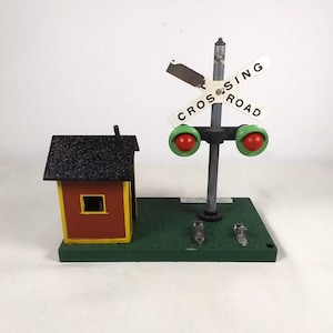 Könnte beinhalten: Ein Spielzeug-Eisenbahn-Überwegsignal mit rotem und grünem Licht, einem kleinen roten und gelben Gebäude und einem Schild mit der Aufschrift "CROSSING ROAD".
