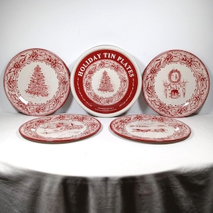 May include: A set of four round tin plates featuring red and white holiday illustrations of Christmas trees, fireplaces, and winter scenes. The plates are displayed with a circular container lid labeled HOLIDAY TIN PLATES ASSORTED SET OF 4.