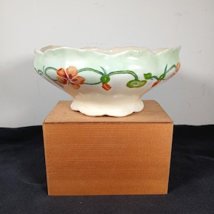 May include: A ceramic bowl with a scalloped rim, painted with orange flowers and green vines against a pale green and cream background. The bowl is set on a small wooden block.