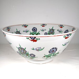 May include: A large, white ceramic bowl with a wide rim and a rounded base. The bowl is decorated with a repeating pattern of blue, green, and red floral designs. The rim is outlined with a thin red band.