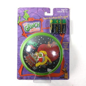 Puede incluir: Un juguete luminoso "Cómo el Grinch robó la Navidad" del Dr. Seuss. La luz verde redonda presenta una escena del Grinch con un saco rojo y un trineo. Incluye cuatro pilas "AA" y es para mayores de 3 años.