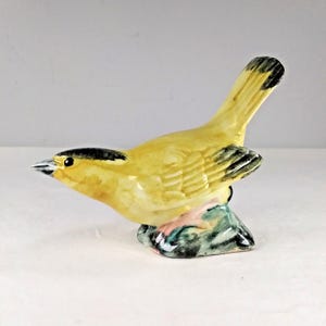May include: A small ceramic figurine of a yellow bird with black markings perched on a green and brown base.