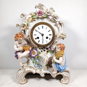 May include: A white porcelain mantel clock featuring gold-trimmed scrollwork, floral accents, and two cherub figures. The clock face has a brass-colored bezel, black Roman numerals, and black hands. One cherub holds a basket, while the other holds wheat.