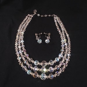 May include: A three-strand iridescent crystal necklace and matching earrings. The necklace is made of clear faceted crystals and has a silver clasp. The earrings are also made of clear faceted crystals and have silver hooks.