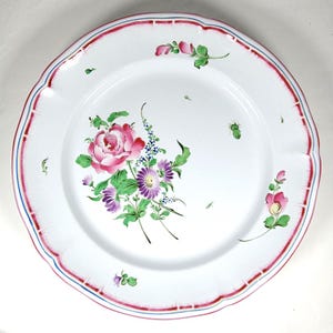 May include: White ceramic plate with a scalloped edge, featuring a hand-painted floral design. The central bouquet includes a large pink rose, purple daisies, and blue accents. The rim has a red and blue border.