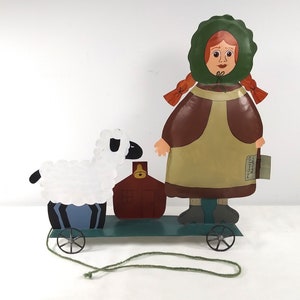 May include: A pull toy featuring a girl in a green hooded dress and a white sheep. The girl is holding a red house with a bell on top. The toy is on wheels and has a green string attached to it.