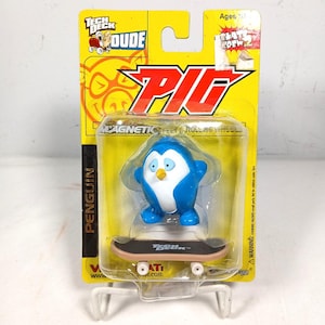 May include: A Tech Deck Dude penguin figure with blue and white colouring and a yellow beak, standing on a black Tech Deck skateboard with white wheels. The toy is encased in a yellow blister pack with the text Tech Deck Dude and Pig.