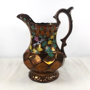 May include: A copper pitcher with a floral design in shades of pink, yellow, green, and white. The pitcher has a textured, diamond-shaped pattern and a handle.