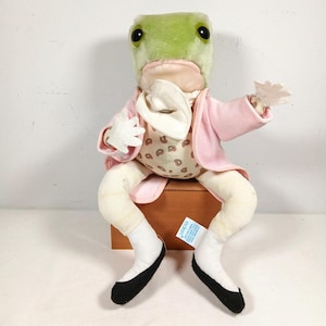 Op de afbeelding: Een pluche kikkerknuffel met grote zwarte ogen, gekleed in een lichtroze jasje over een crèmekleurig vest met een bruin paisley-patroon. De kikker draagt een witte cravat, witte handschoenen, witte sokken en zwarte schoenen met een blauw label.