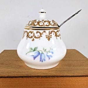 Może przedstawiać: Biała cukiernica z porcelany z pozłacanym wykończeniem i kwiatowym wzorem. Pokrywka ma złoty guzik, a cukiernica jest wyposażona w małą srebrną łyżeczkę.