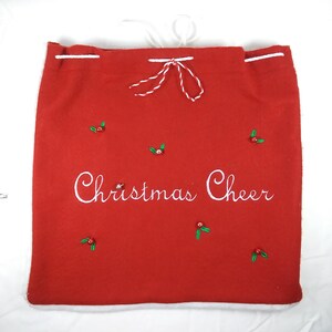 May include: Red felt Christmas gift bag with white drawstring and white embroidery that says "Christmas Cheer". The bag is decorated with green holly leaves and red berries.