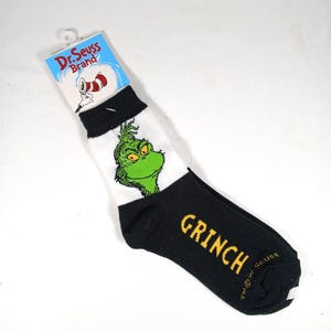 以下が含まれることがあります： Grinchのキャラクターが描かれた、黒、白、緑の靴下のペア。靴下の黒い部分には黄色で「GRINCH」の文字が書かれています。Dr. Seuss Brandのロゴが付属のタグに表示されています。