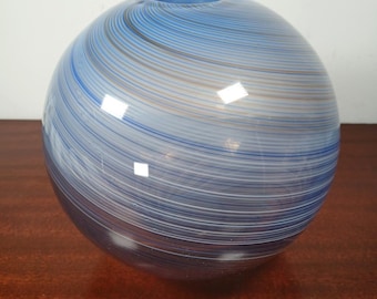 May include: A spherical glass vase with a blue and white striped pattern. The vase has a small opening at the top and is sitting on a wooden surface. The stripes are in shades of blue, white, and a hint of brown.