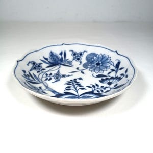 May include: A white ceramic plate with a scalloped edge, adorned with a blue floral pattern. The design features stylized flowers, leaves, and branches, creating a classic, elegant aesthetic. The plate is round and appears to be approximately 6 inches in diameter.