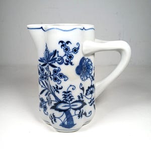 Puede incluir: Jarra de cerámica blanca con un estampado floral azul. La jarra tiene un borde festoneado y un asa curva. El diseño floral azul incluye flores, hojas y tallos. La jarra mide aproximadamente 15 cm de altura.