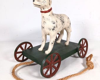 Könnte beinhalten: Eine weiße Hundefigur mit schwarzen Flecken und einem roten Halsband, montiert auf einer dunkelgrünen Holzplattform mit vier roten Rädern. Ein dickes, beigefarbenes Juteseil ist an der Vorderseite befestigt, um das Spielzeug zu ziehen.