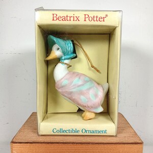 May include: A Beatrix Potter collectible ornament featuring a white duck wearing a light blue bonnet and a pink shawl with light blue spots. The duck is inside a rectangular box with the text Beatrix Potter and Collectible Ornament printed.