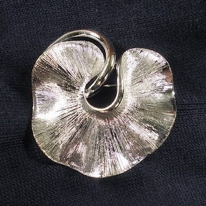 Puede incluir: Un broche plateado en forma de hoja estilizada con un tallo retorcido. La hoja tiene una superficie texturizada.