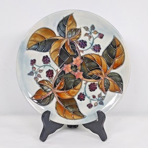 May include: A white ceramic plate with a decorative design of blackberries and leaves in shades of green, brown, and gold.