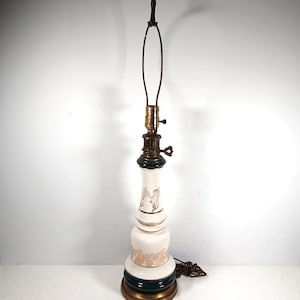 May include: A table lamp with a white ceramic base featuring gold floral patterns and a dark green band near the bottom. The lamp has a brass-colored metal base, a brass socket with a turn-knob switch, and a metal harp attached to the top fixture.