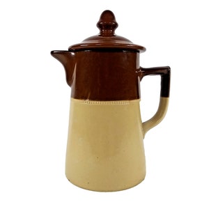 May include: A brown and beige ceramic pitcher with a lid and a handle. The pitcher has a decorative band around the middle.